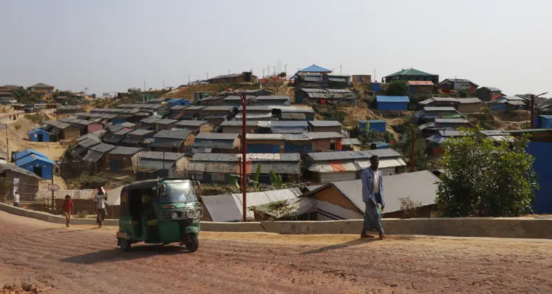 Rows of shelters with blue and grey roofs on a hillside in a Cox's Bazar camp, a man walking and an auto-rickshaw on a brick road